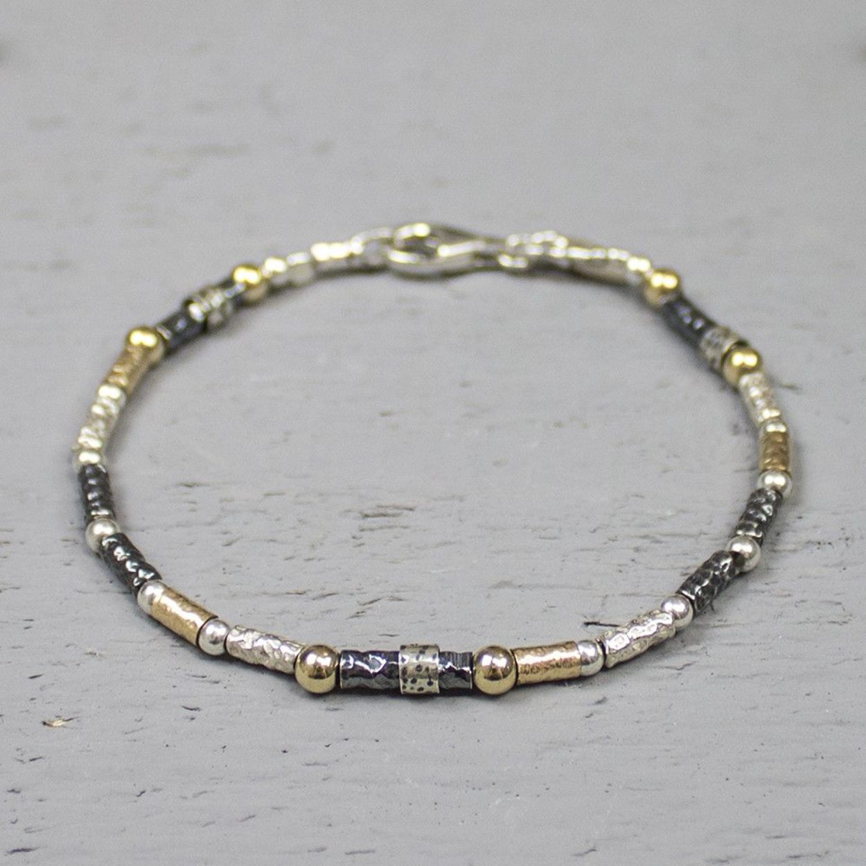 3 kleurige fantasiearmband, met de karakteristieke kleuren van J&eacute;h Jewels: zilver, geoxideerd zilver en goldfilled. 
De armband bestaat uit kleine buisjes/stukjes die aan mekaar geregen zijn. 
Hij is 19.5 cm lang en 4 mm breed.